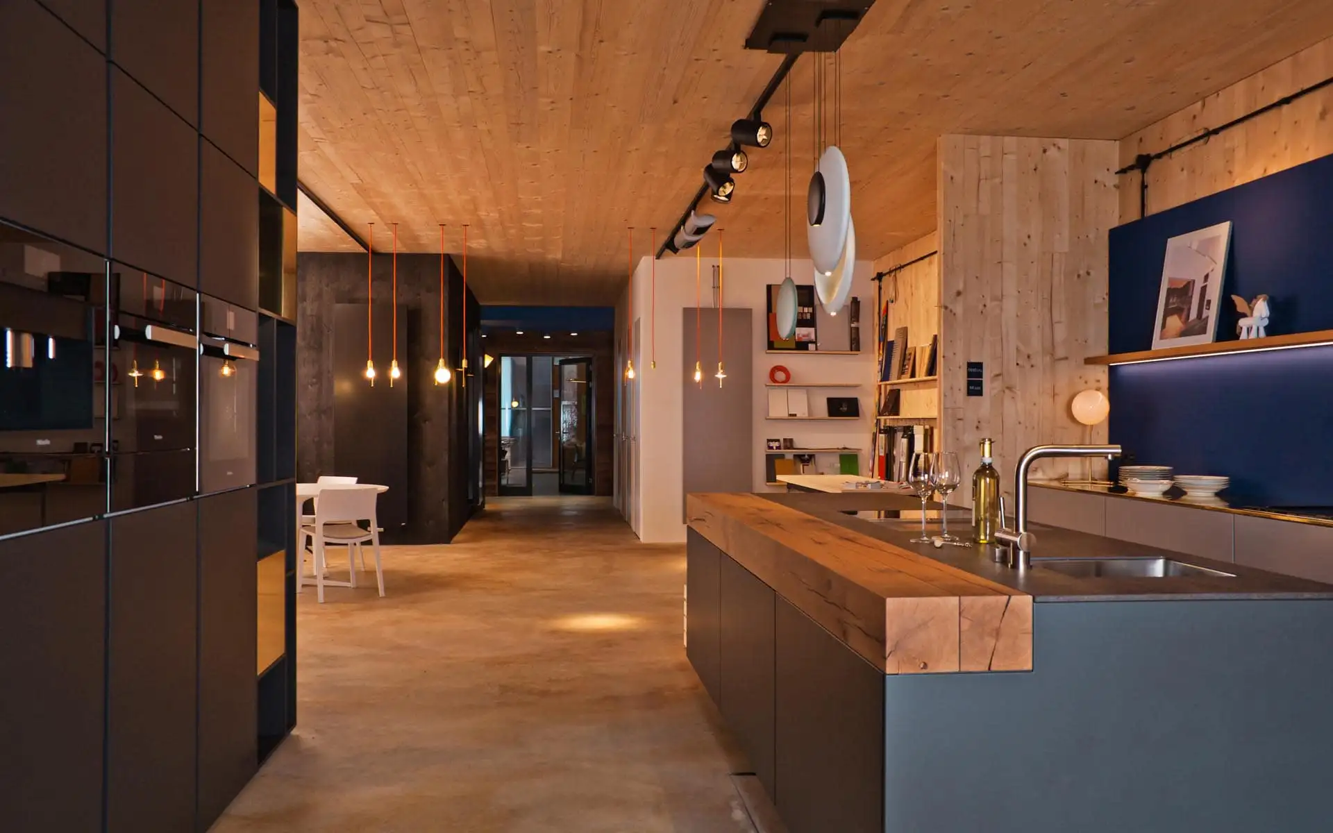 Innenraum mit Kücheninsel aus dunklem Material, massiver Holzarbeitsplatte und deckenintegrierten Leuchten.