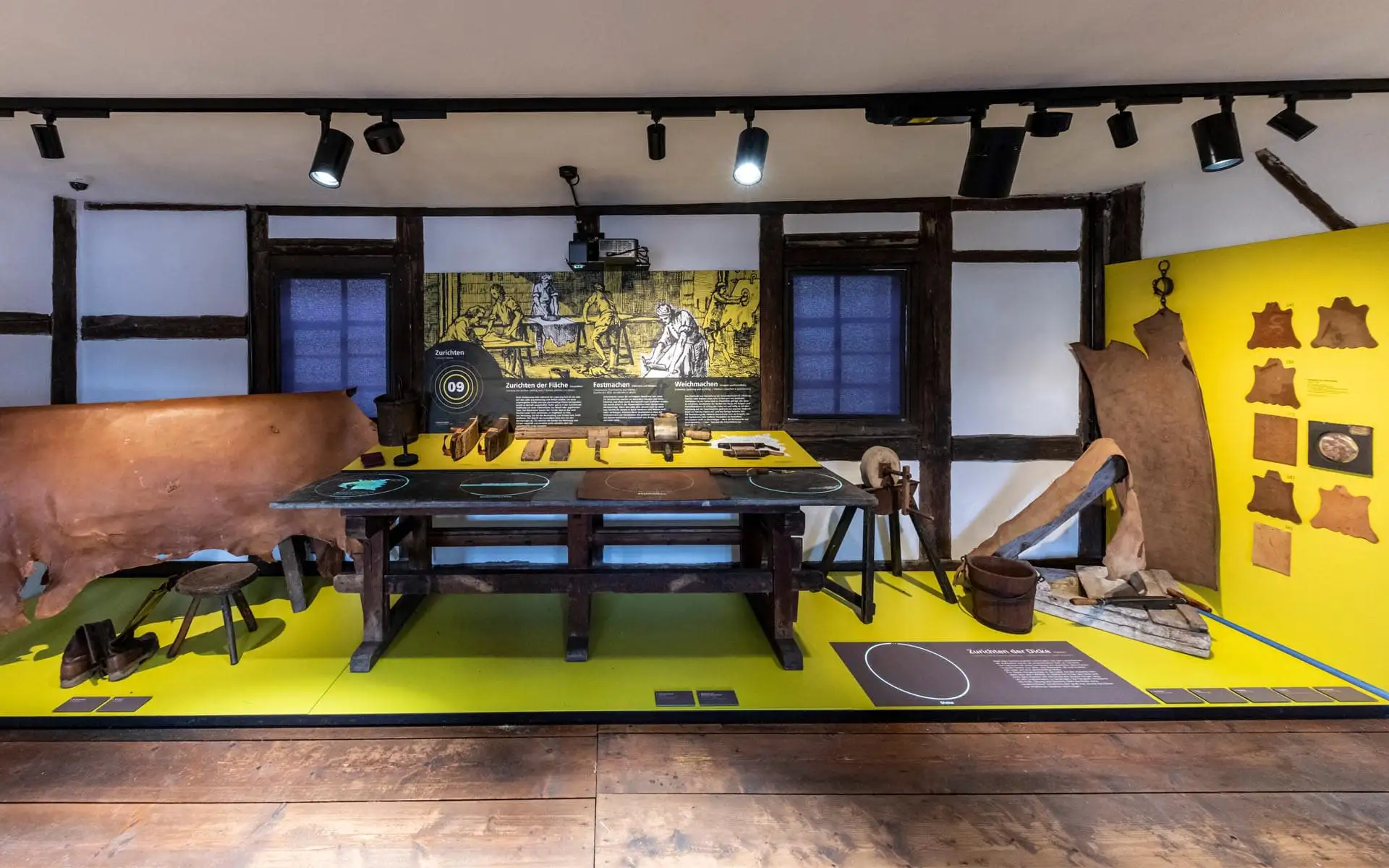 Gerberwerkstatt: Werkbank mit Werkzeugen, gelbe Tafeln, historische Zeichnung im Hintergrund