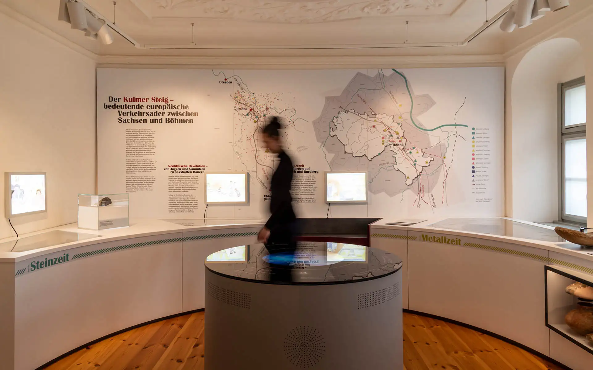 Ausstellungsraum mit gebogener Vitrinenzeile, runder Medienstation und großem Wandplan 'Kulmer Steig'.