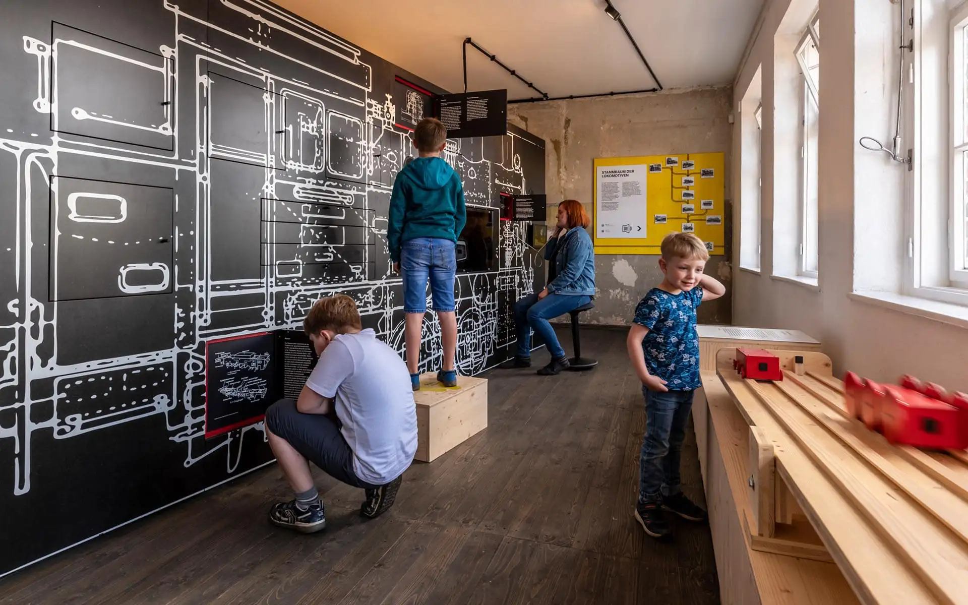 Großer Ausstellungsraum mit Holzkonstruktionen, Leinwandprojektion und Besuchern