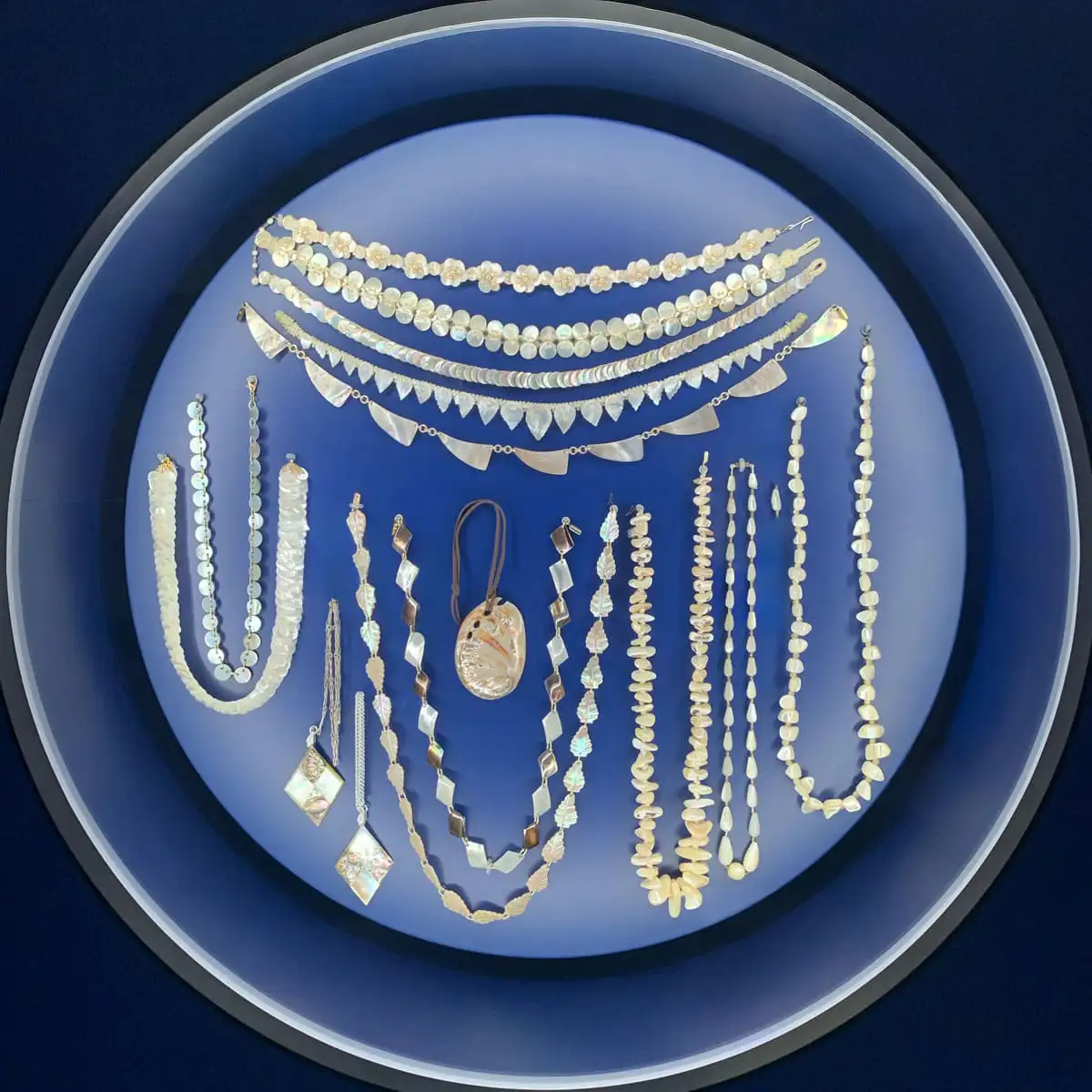 Runde Vitrine mit Perlmutterschmuck, darunter Ketten, Anhänger und Ohrringe, symmetrisch auf blauem Hintergrund arrangiert.