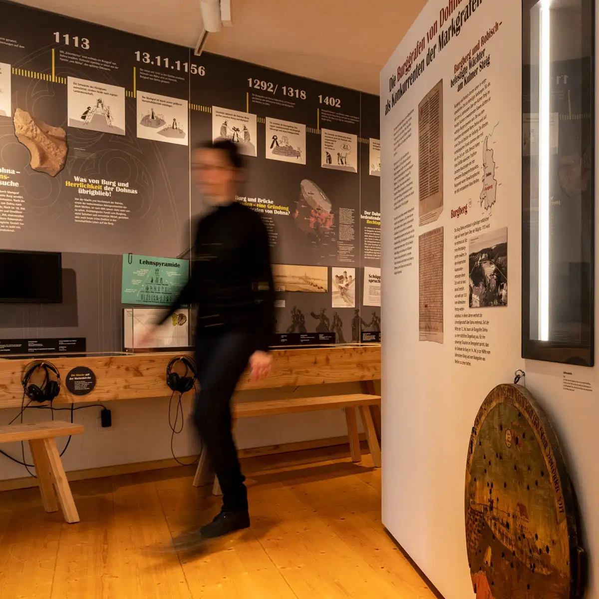 Detail der Zeitstrahl-Wand. Besucherin geht an Infotafeln und Sitzbänken vorbei.