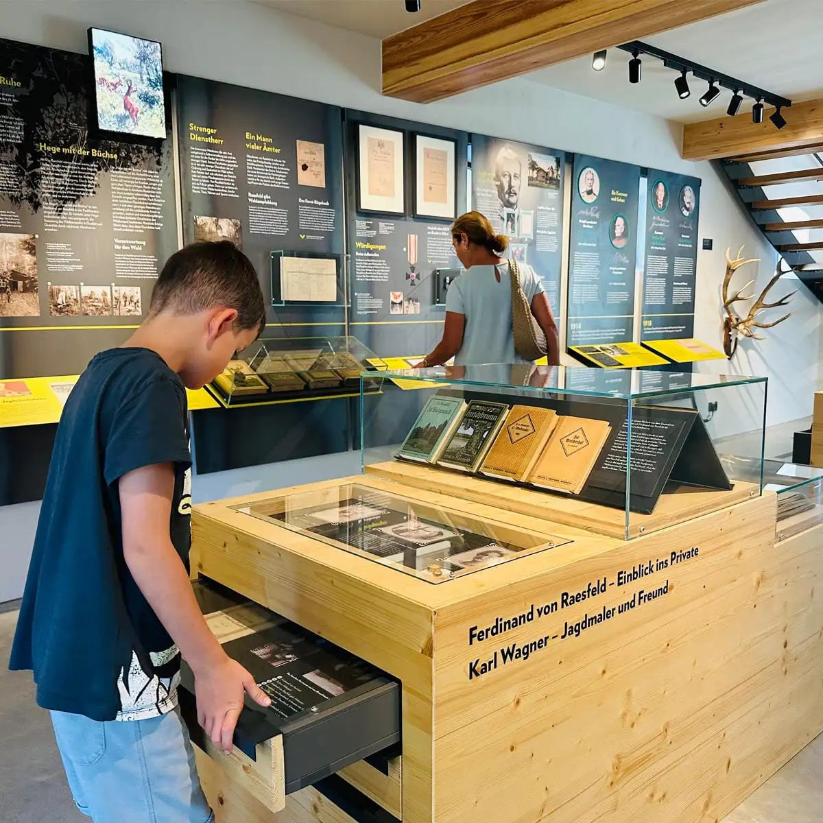 Junge zieht eine Schublade einer Vitrine auf. Im Hintergrund weitere Exponate und Wandgrafiken, daneben eine Besucherin.