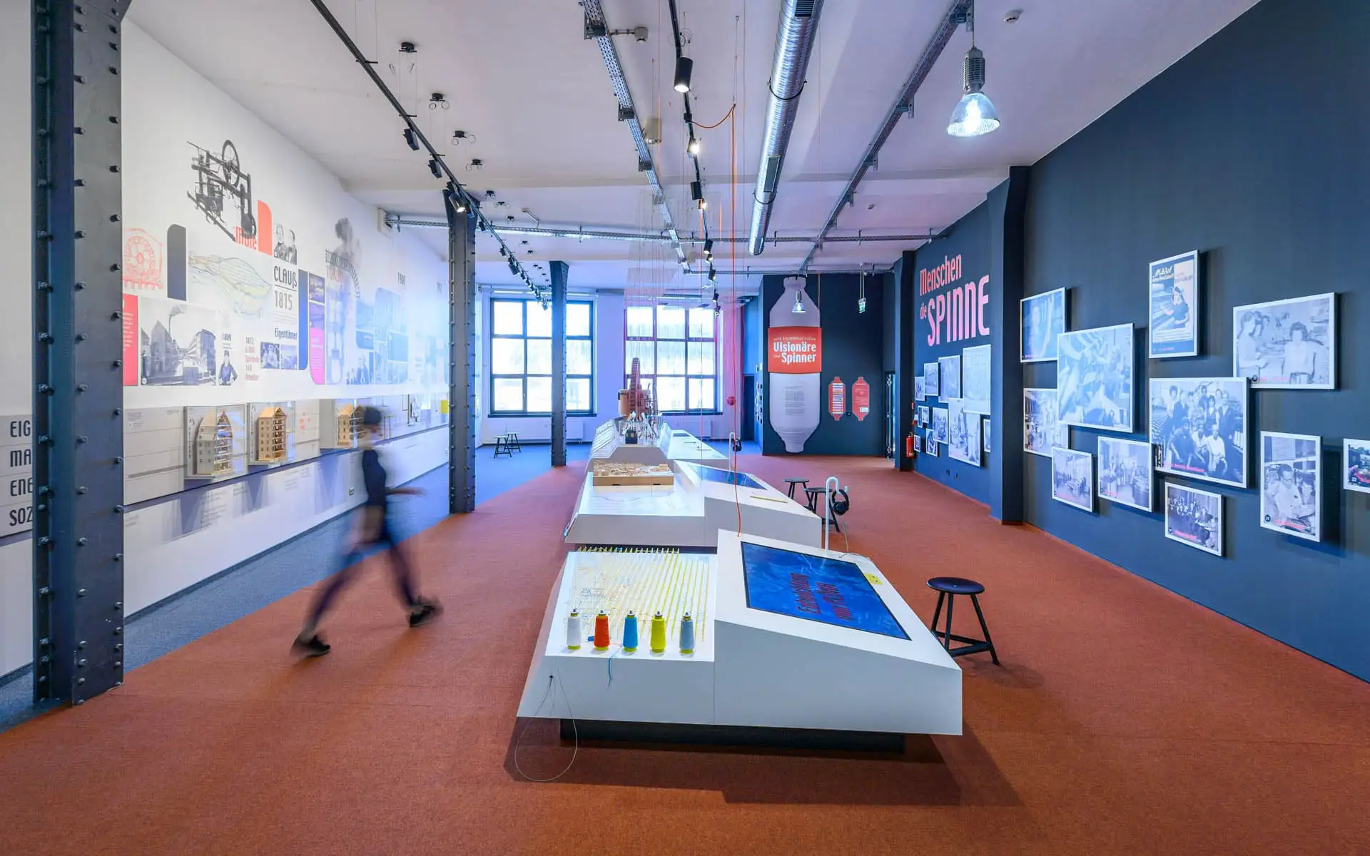 Blick durch den Ausstellungsraum mit zentraler Präsentationsfläche, Fotowand und Vitrinen