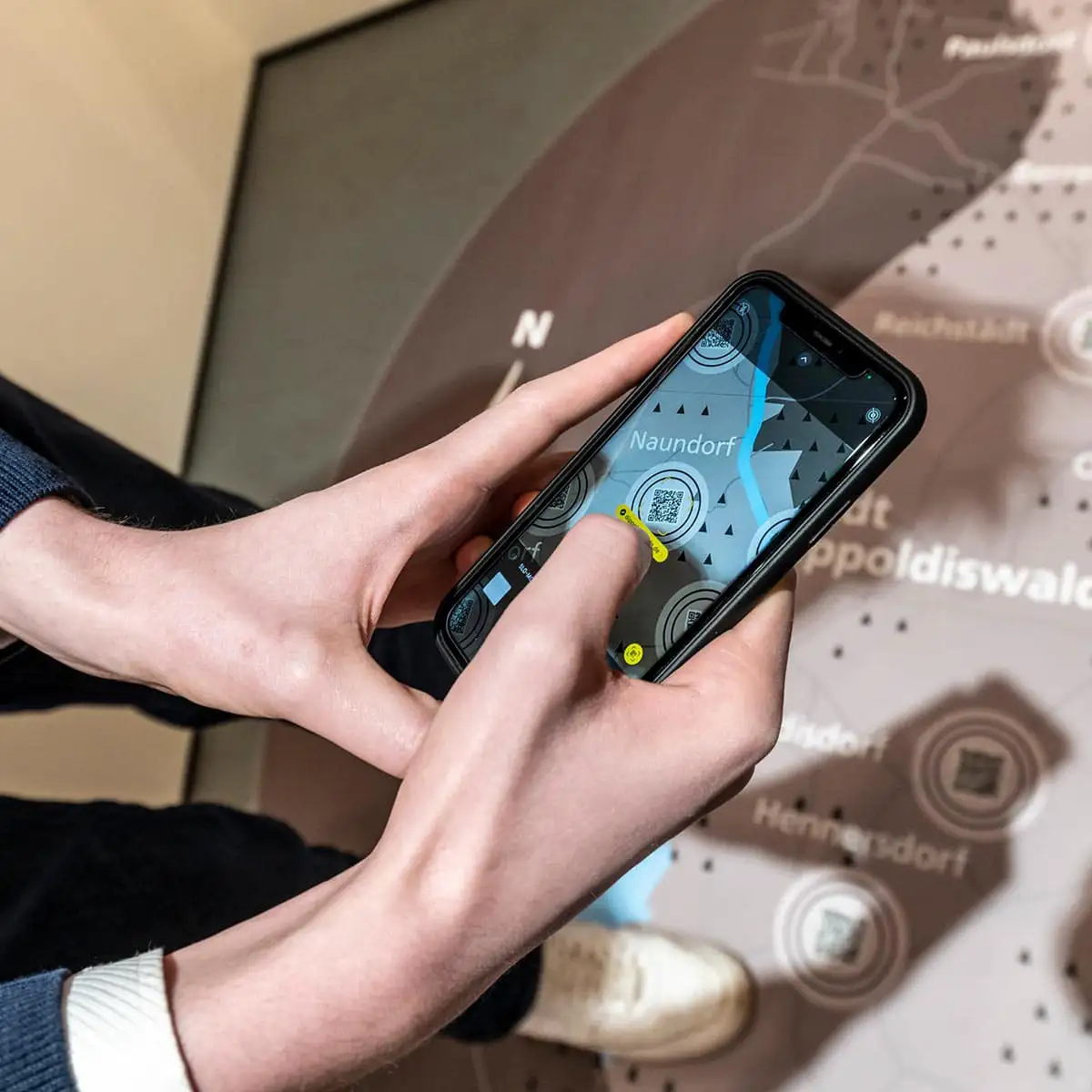Hände bedienen ein Smartphone mit Ausstellungs-App vor einer Bodenkarte, Ortsname 'Naundorf' ist zu sehen