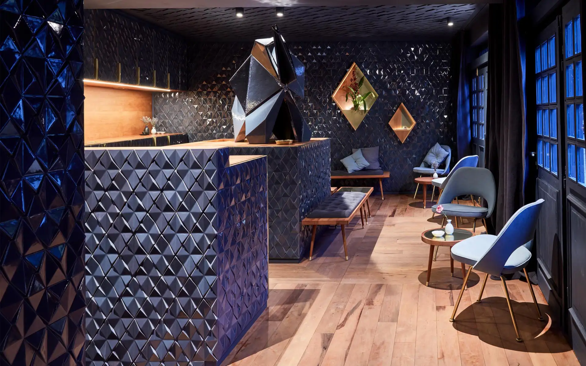 Dunkle Bar mit glasierten, geometrischen Wandfliesen, Holzboden und modernem Sitzbereich in Blautönen.