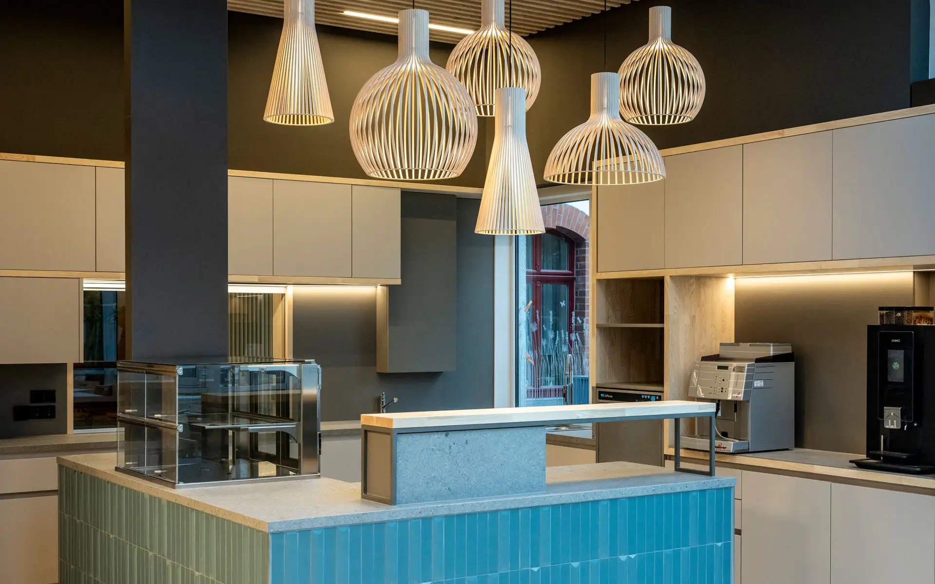 Moderne Cafétheke mit türkisfarbener Fliesenfront, heller Arbeitsfläche und skulpturalen Pendelleuchten aus Holz.