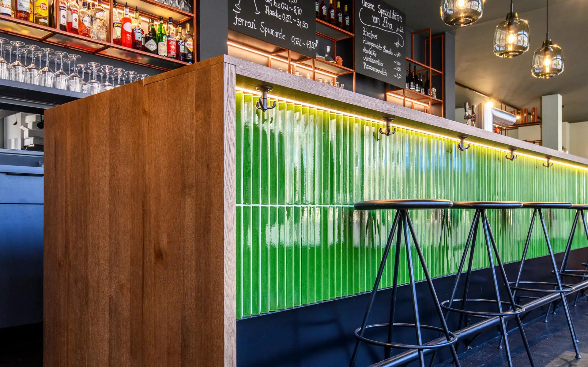 Nahaufnahme einer grünen, vertikal gekachelten Bartheke mit Holzabschluss und schwarzen Barhockern davor.