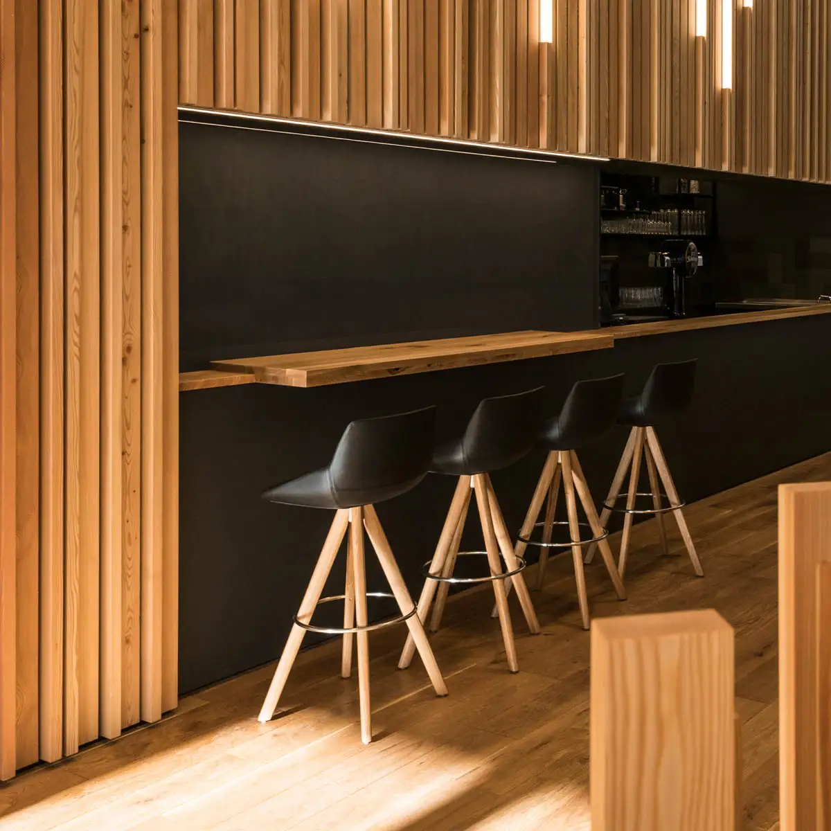 Moderne Bar mit hohen Holzpaneelen, schwarzer Theke und Barhockern aus Holz und Leder.