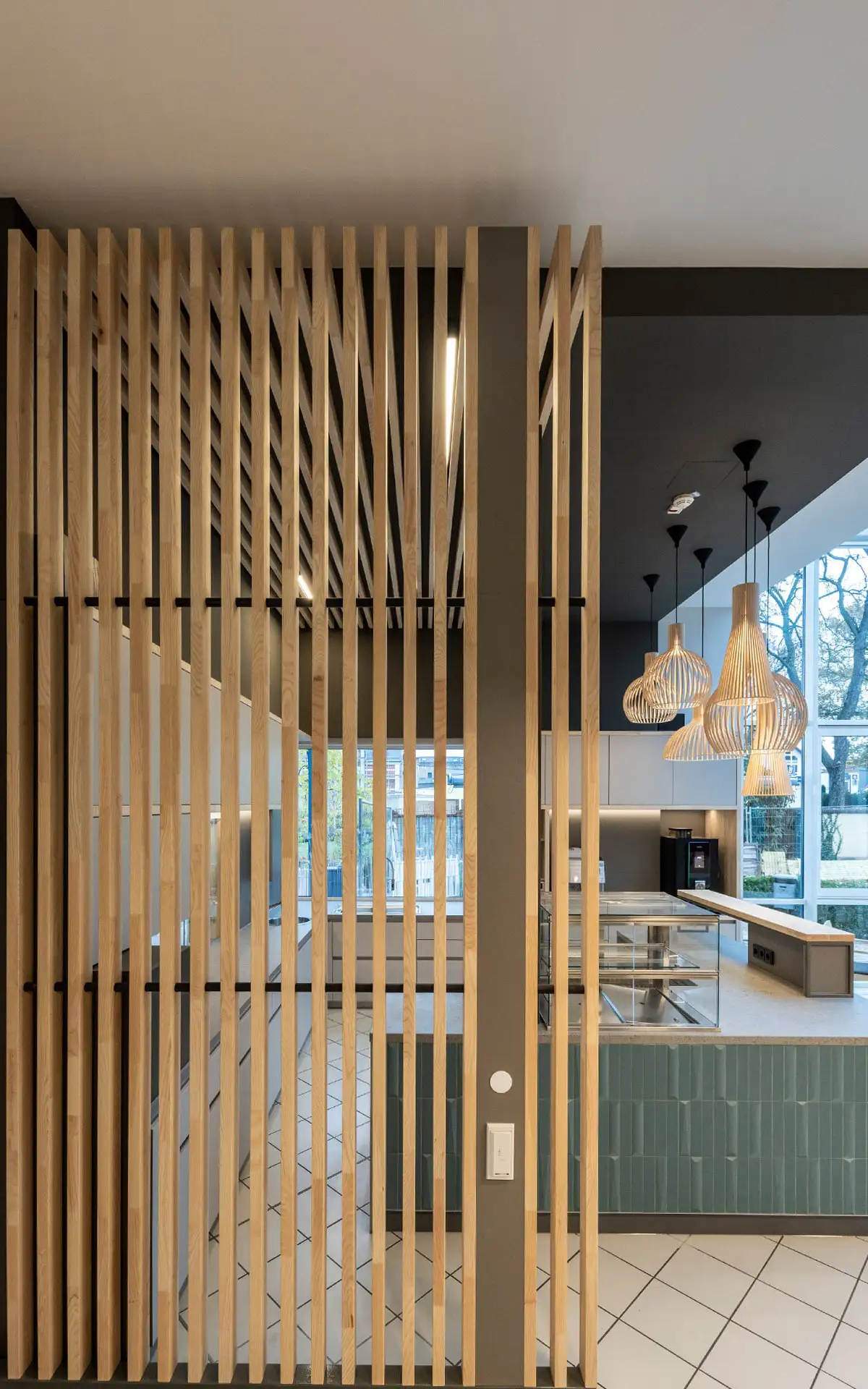 Vertikale Holzlamellen als transparenter Raumteiler zwischen Gastraum und Thekenbereich mit integrierter Beleuchtung.