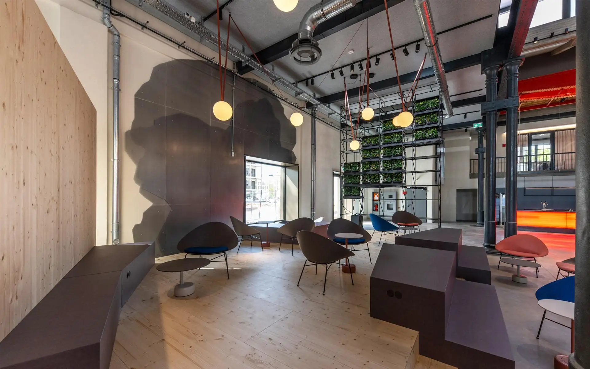 Offener Aufenthaltsbereich in industrieller Architektur mit farbigen Lounge-Sesseln, Pflanzenwand und runden Leuchten.