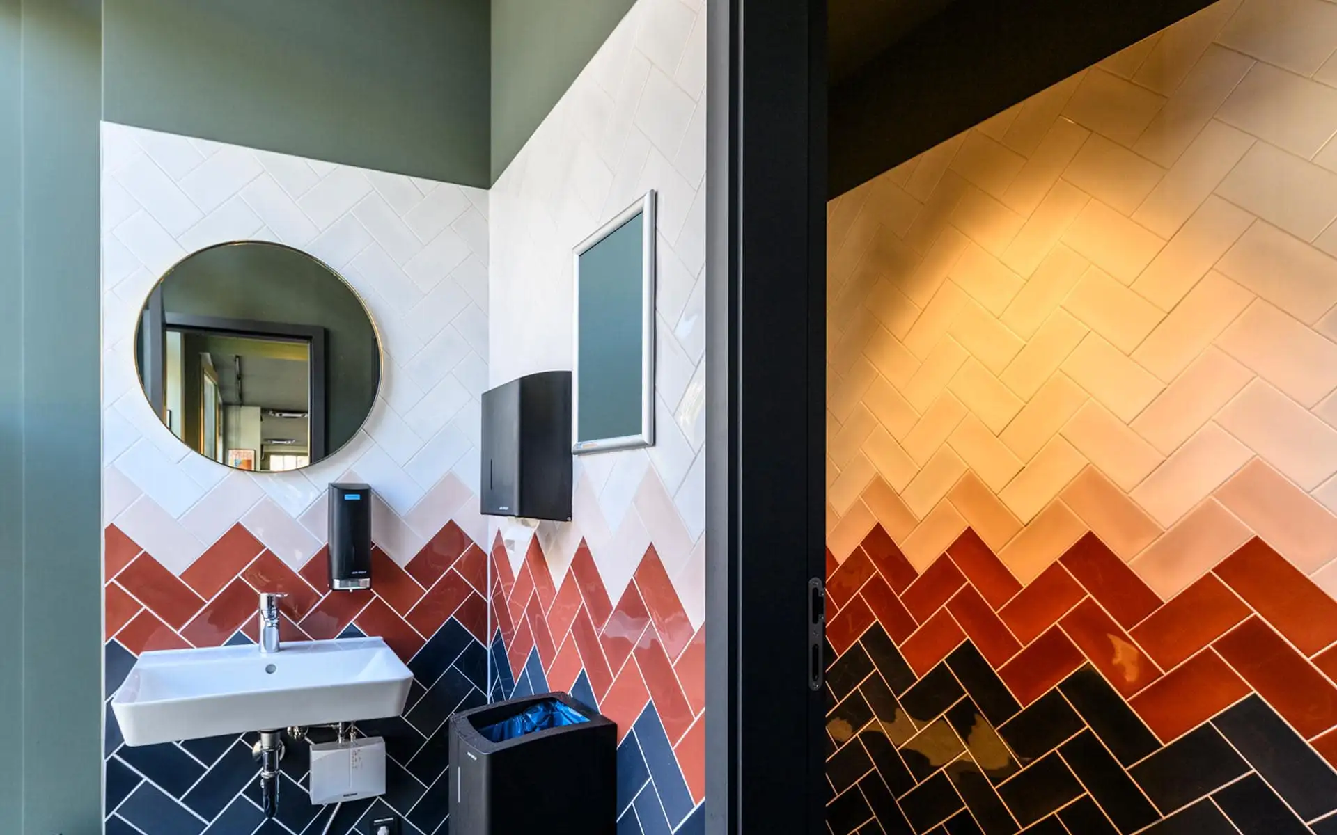 Waschraum mit bunten, diagonal verlegten Wandfliesen in Weiß, Rot und Blau, rundem Spiegel und Blick in den Duschbereich.