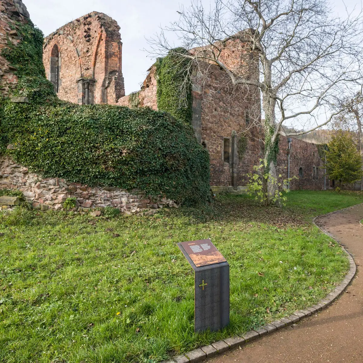 Seitenansicht der überwachsenen Kirchenruine mit Baum und Weg, davor eine Informationstafel