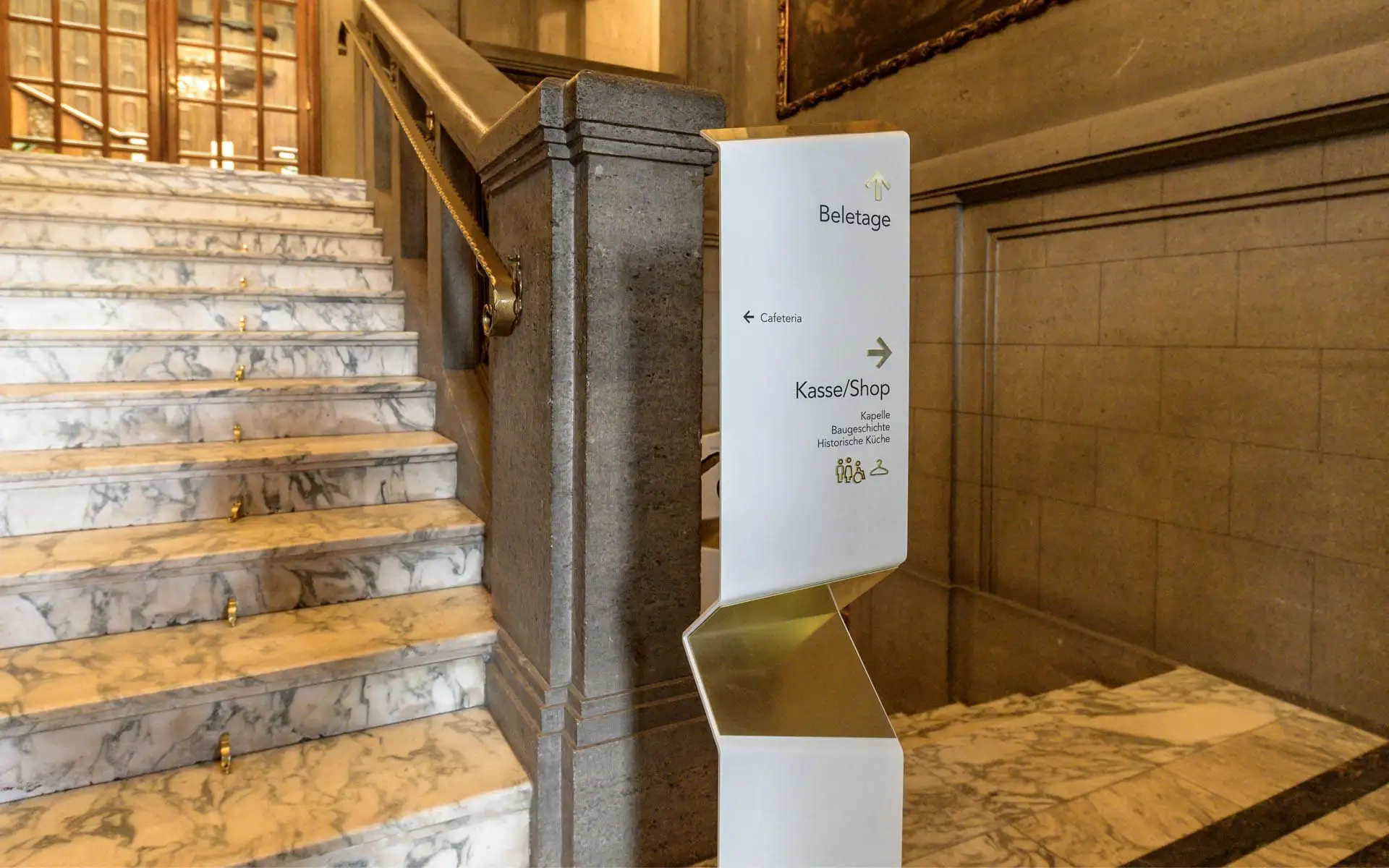 Leitsystem-Schild am Treppenaufgang im Schloss Waldenburg mit Marmorstufen und historischer Atmosphäre.