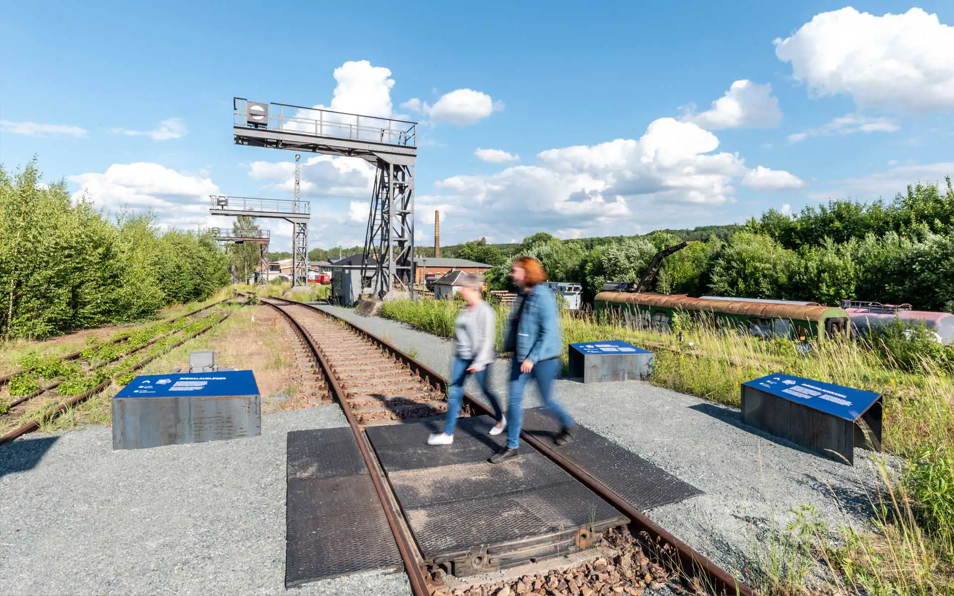 Bahnübergang und Informationsinseln im Freigelände eines Eisenbahnmuseums mit zwei vorbeigehenden Personen.