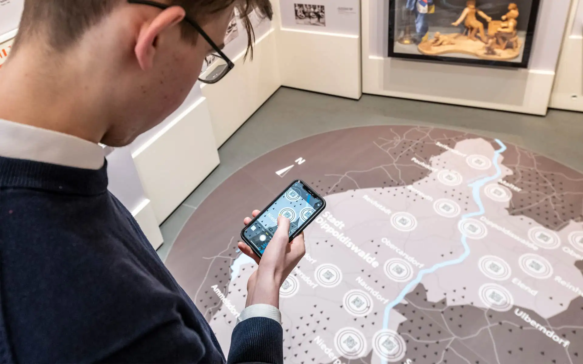 Besucher nutzt ein Smartphone über einer großen Bodenkarte in einer interaktiven Ausstellung.