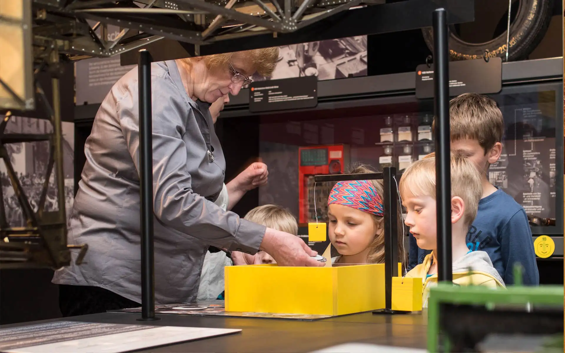 Museumsführerin zeigt Kindern ein Exponat an einer gelben Mitmachstation.