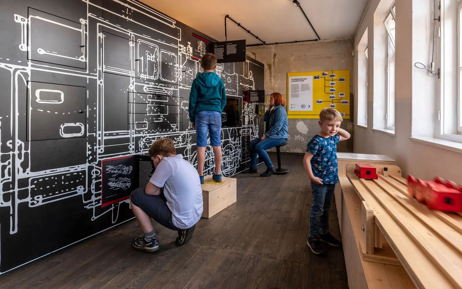 Kinder erkunden einen Ausstellungsraum mit großer Lokomotivzeichnung und Modellbahnstation.