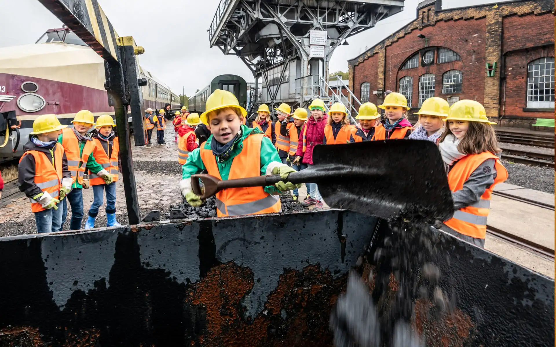 Kinder in Warnwesten und Helmen schaufeln Kohle auf einem Eisenbahngelände.