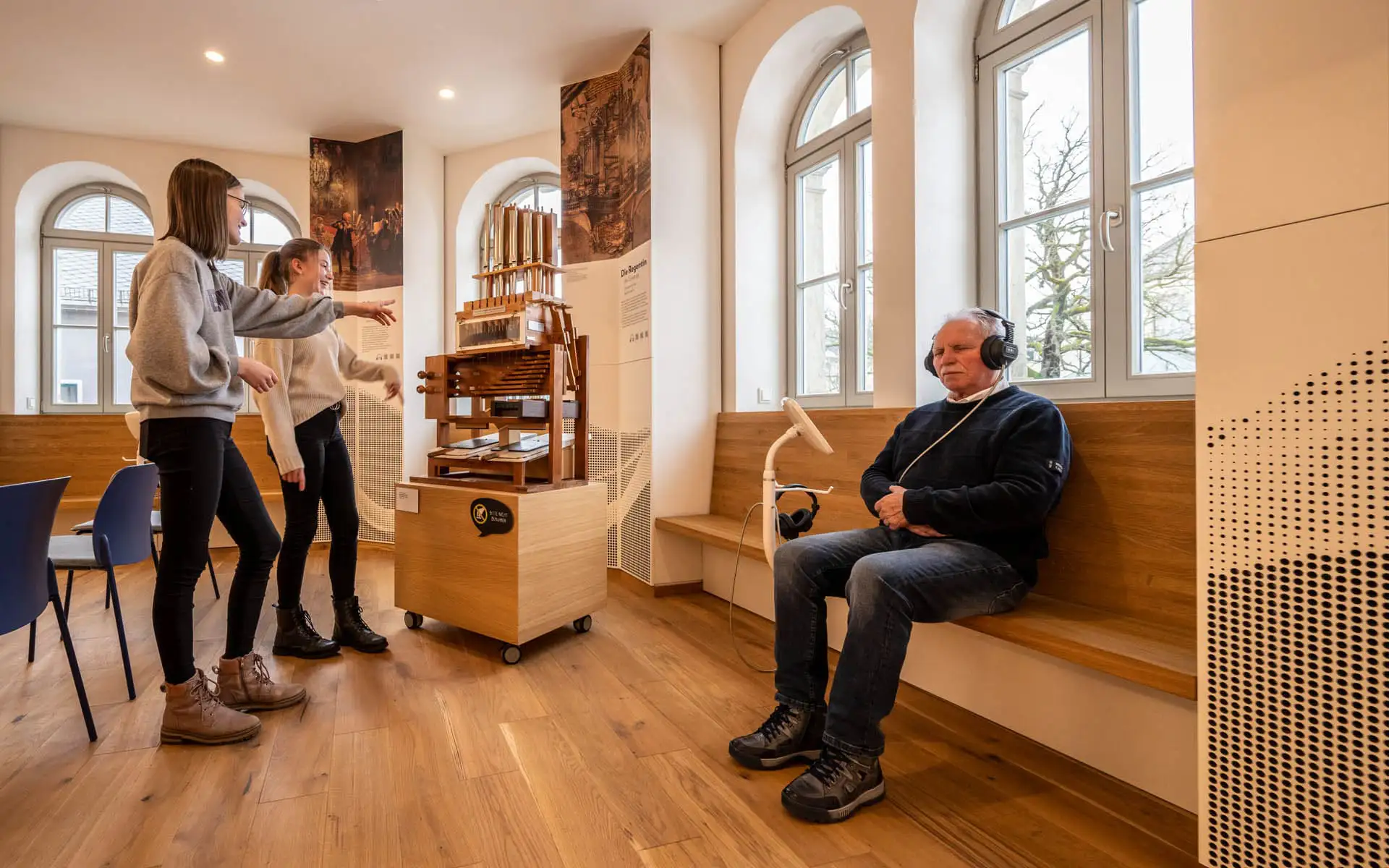 Mann mit Kopfhörern sitzt an einer Hörstation, während zwei Jugendliche eine Museumsstation betrachten.