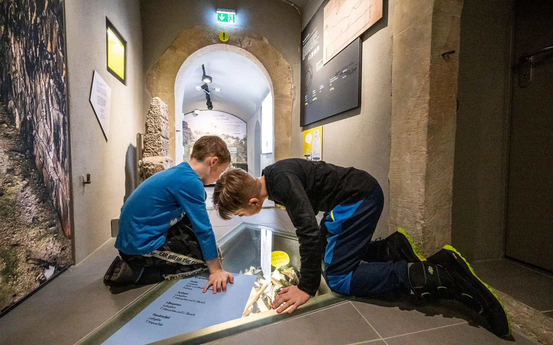 Zwei Jungen knien über einer bodentiefen Vitrine in einem schmalen Ausstellungsraum.