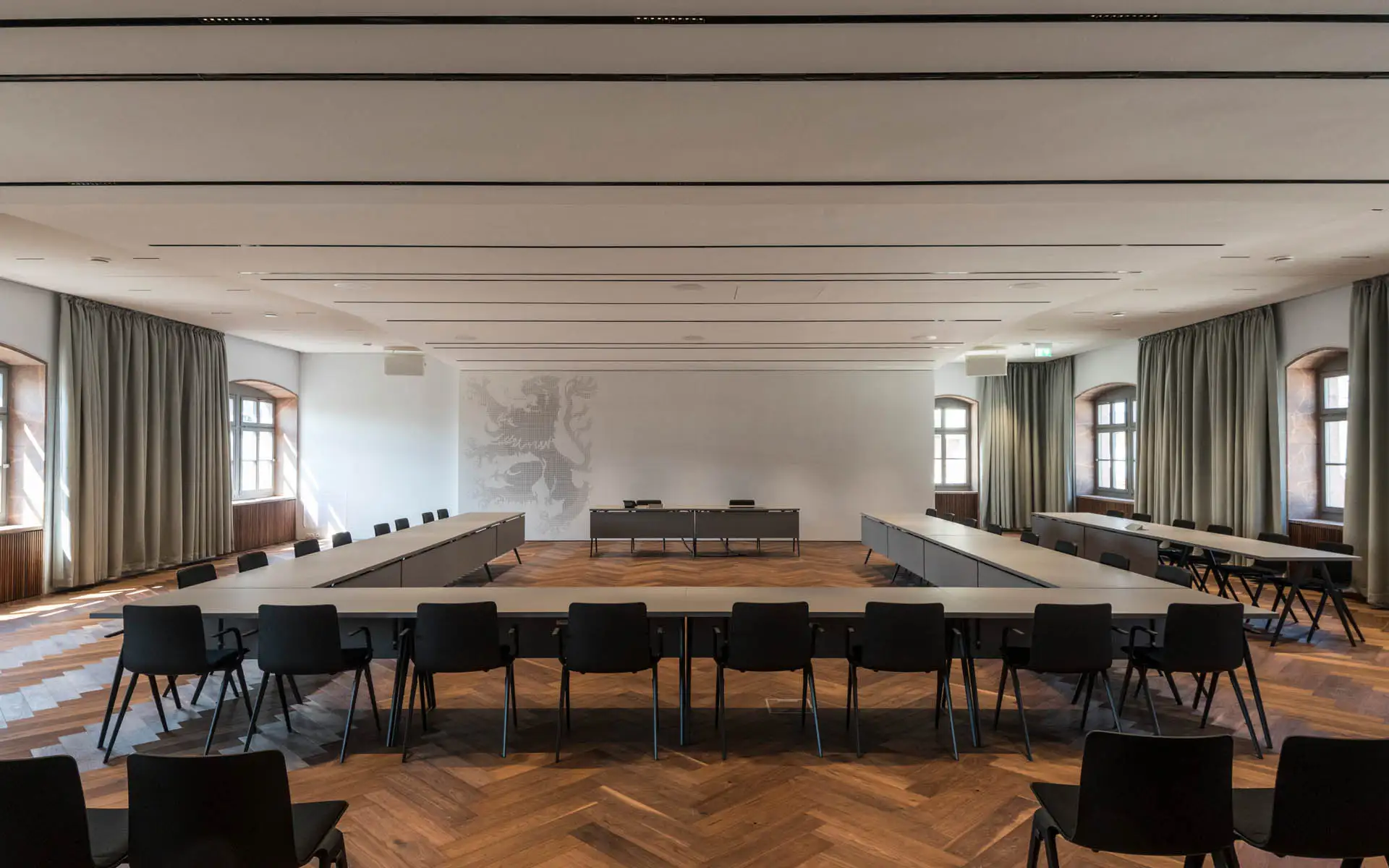 Blick zur Front des Saals: Podium, akustisch gegliederte Decke, hohe Fenster mit Vorhängen