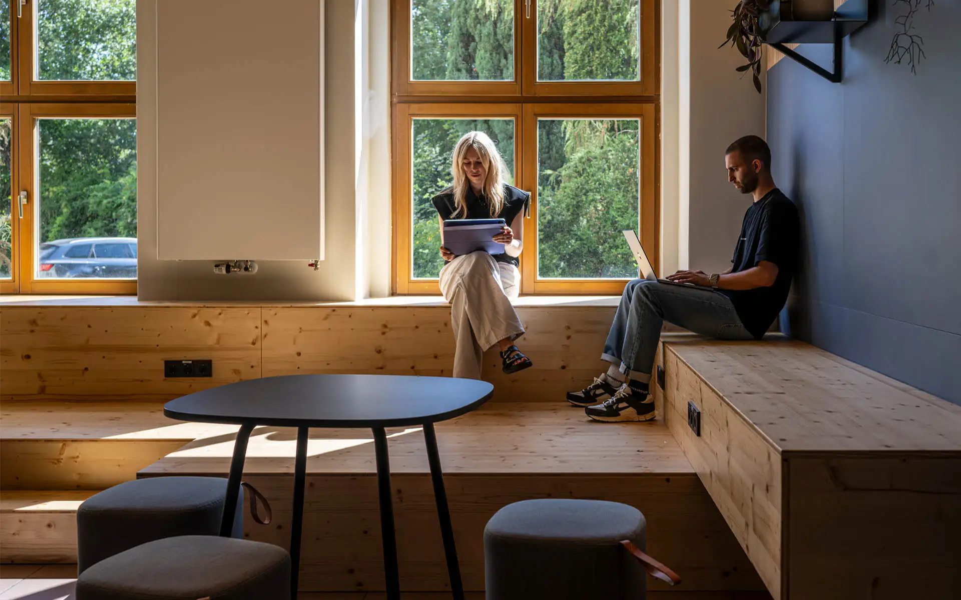 Sitzstufen aus Holz vor großen Fenstern, eine Person liest, eine arbeitet am Laptop.