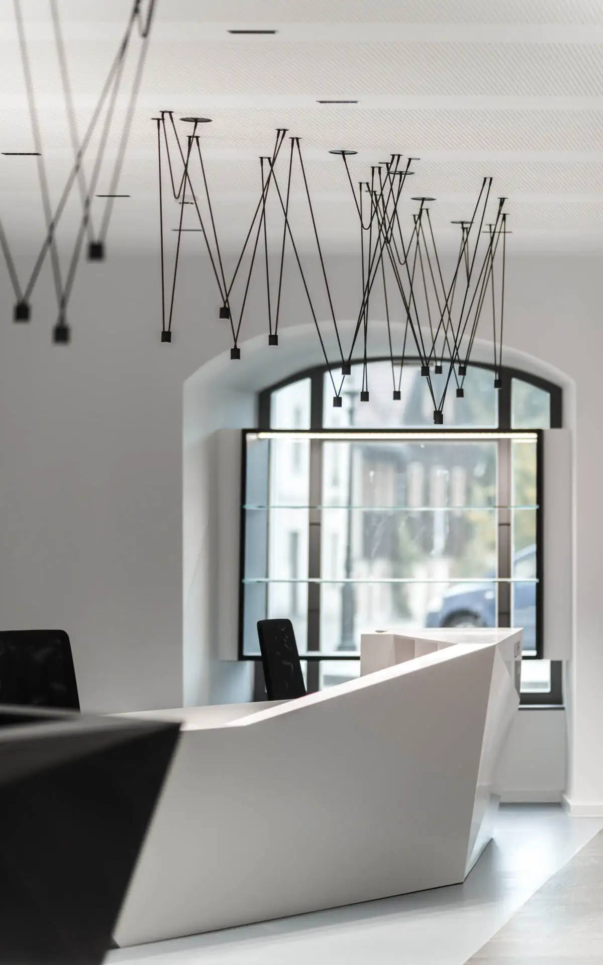 Blick entlang des Empfangstresens auf ein großes Fenster, filigrane Pendelleuchten zeichnen Z-Linien an der Decke