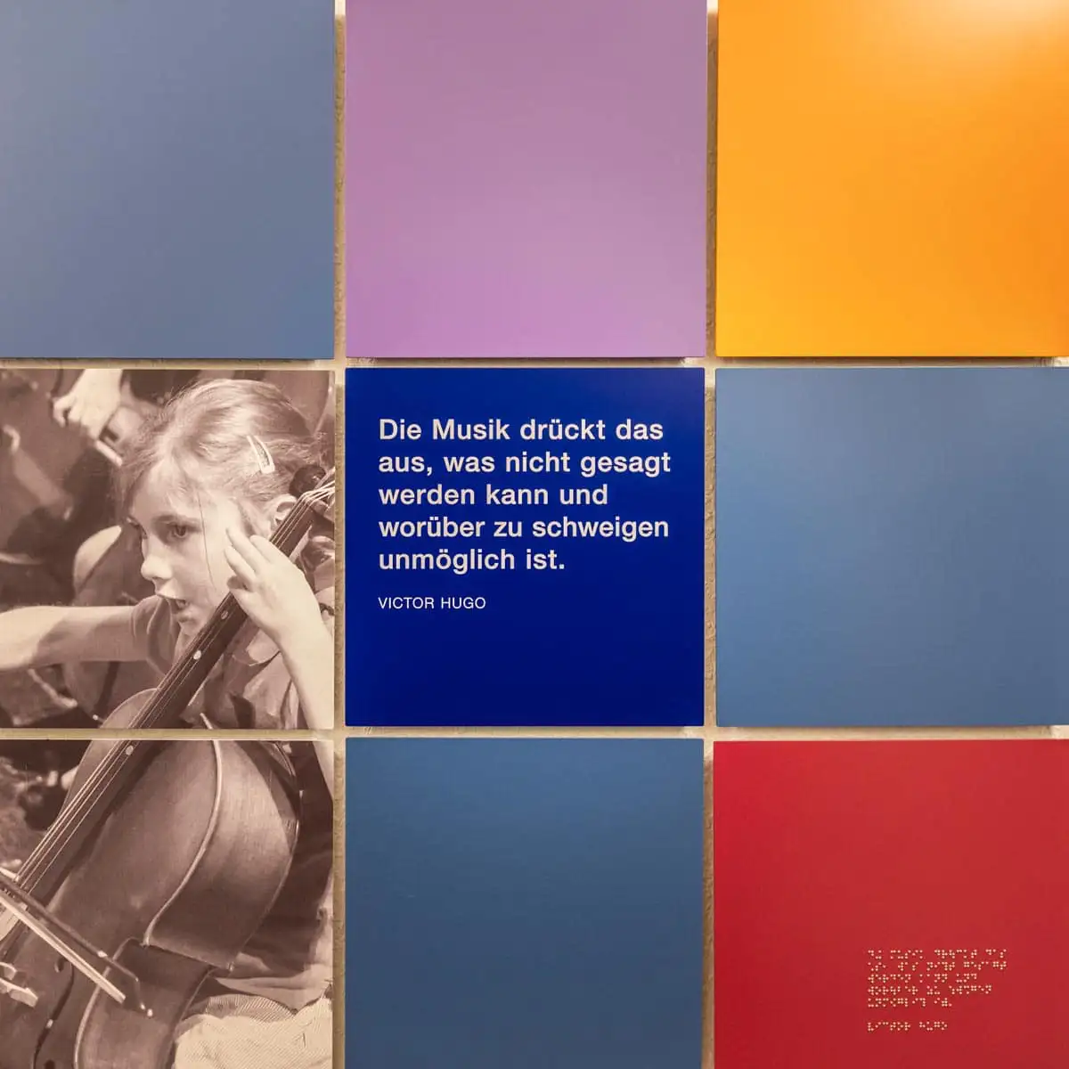 Mosaik aus farbigen Wandpaneelen mit Zitat von Victor Hugo und Foto eines Kindes mit Cello.