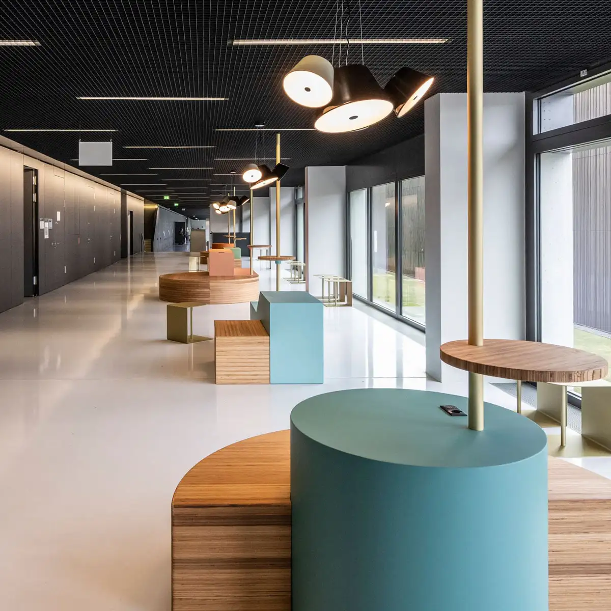 Langer Korridor mit mehreren Sitzinseln in Pastellfarben, große Fenster rechts, schwarze Decke mit Leuchten