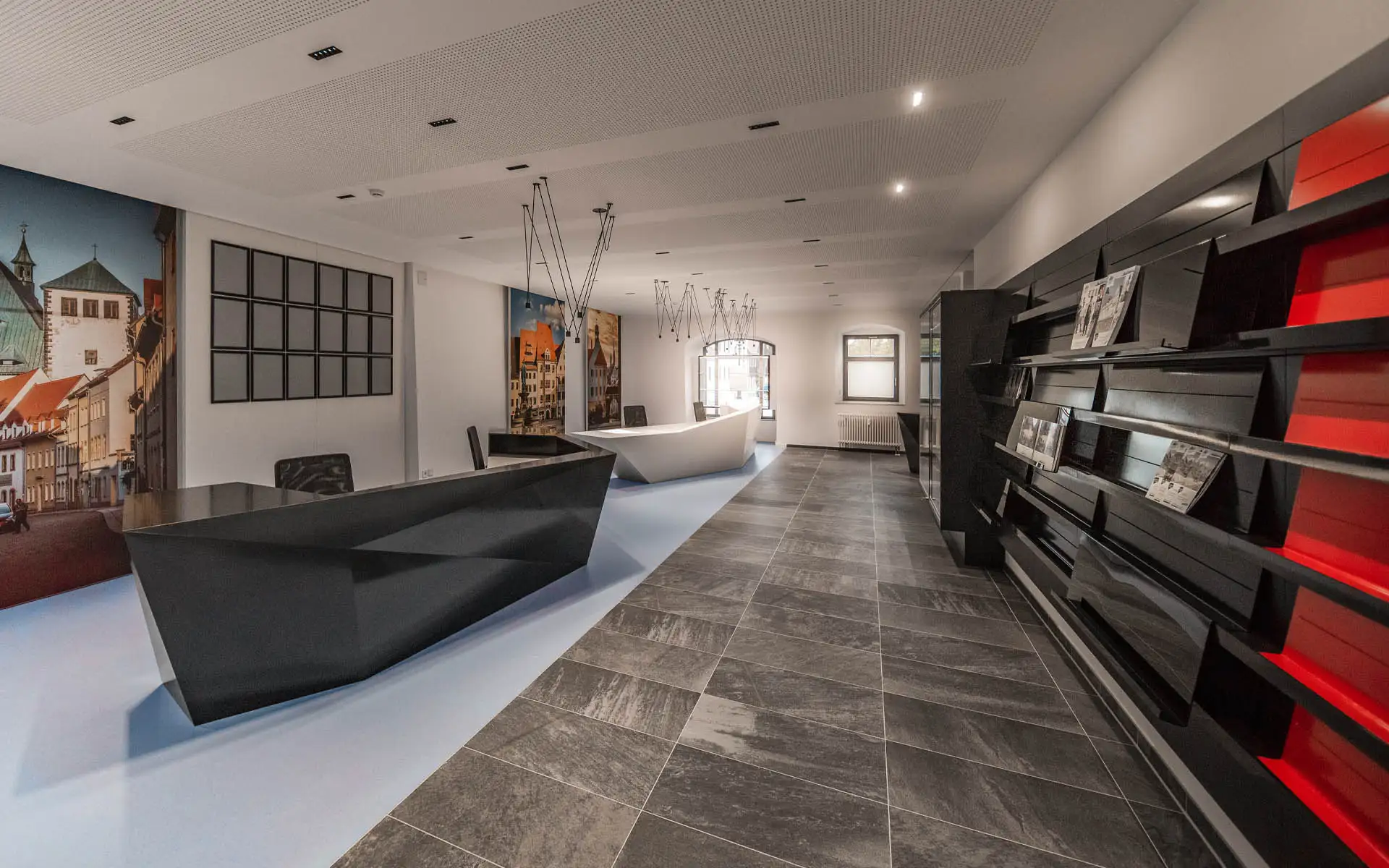 Langer Raum mit schwarzem Counter links, weißem Empfangstresen und Wandregal für Drucksachen rechts