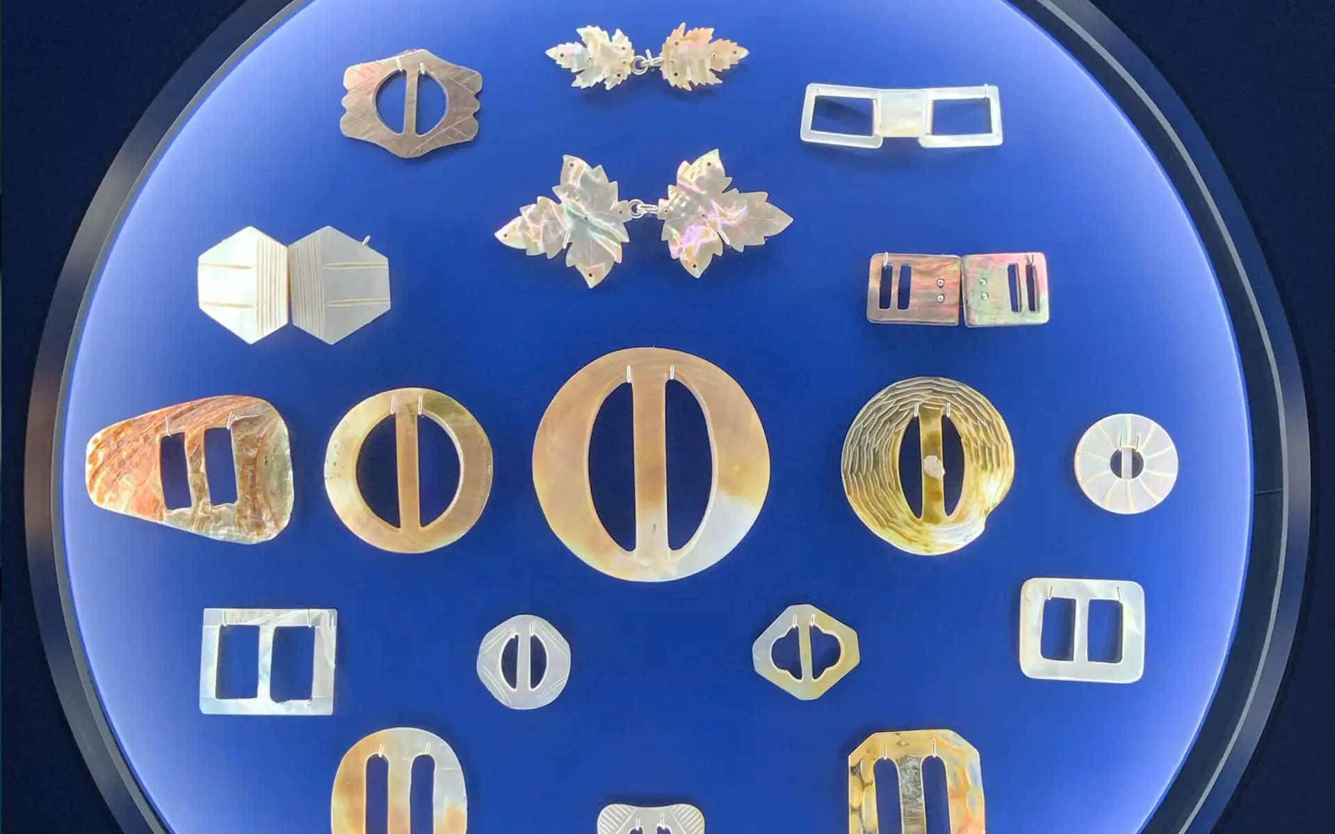 Kreisförmig angeordnete Perlmuttobjekte wie Schnallen und Zierelemente auf blauem Ausstellungshintergrund.