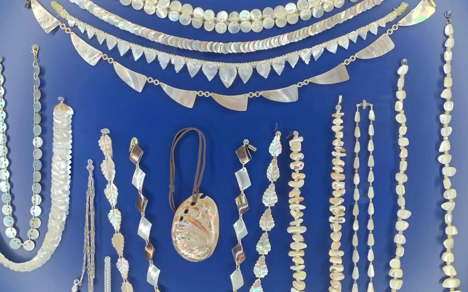 Historischer Perlmuttschmuck mit Ketten, Anhängern und Ohrschmuck, geordnet auf blauem Textilgrund.