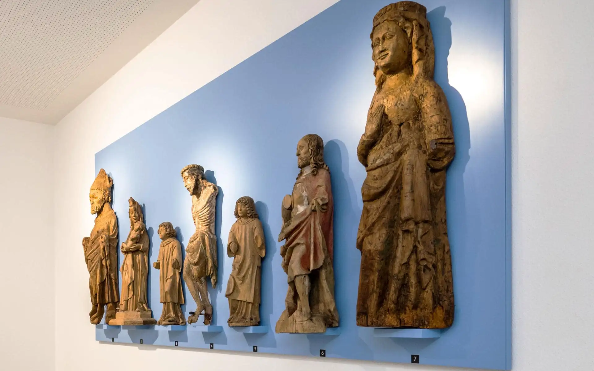 Reihe historischer sakraler Holzskulpturen an einer hellblauen Wand, einzeln beleuchtet und nummeriert.