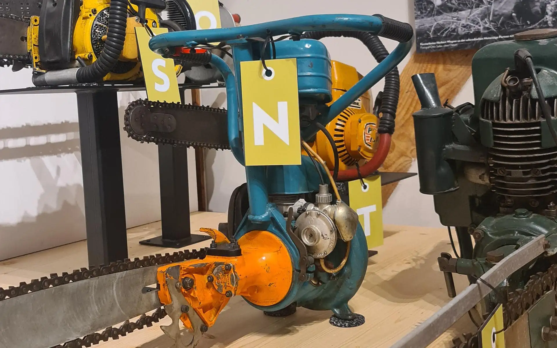 Historische Motorsäge mit blauem Gehäuse und orangefarbenem Schneidkopf als Museumsobjekt.