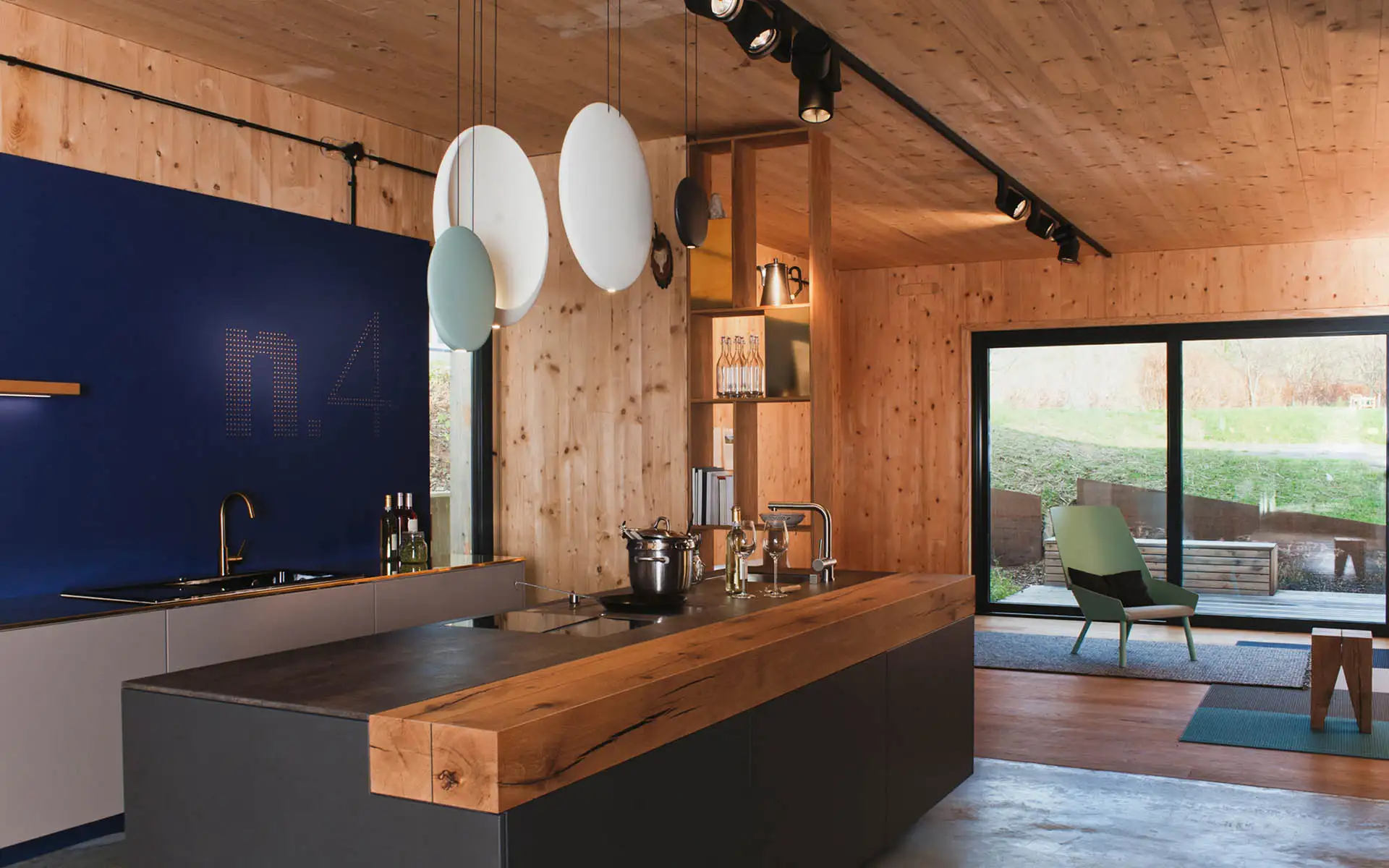 Moderne Wohnküche mit massiver Holzarbeitsplatte, dunkler Kücheninsel, blauer Akzentwand und runden Pendelleuchten aus Glas.