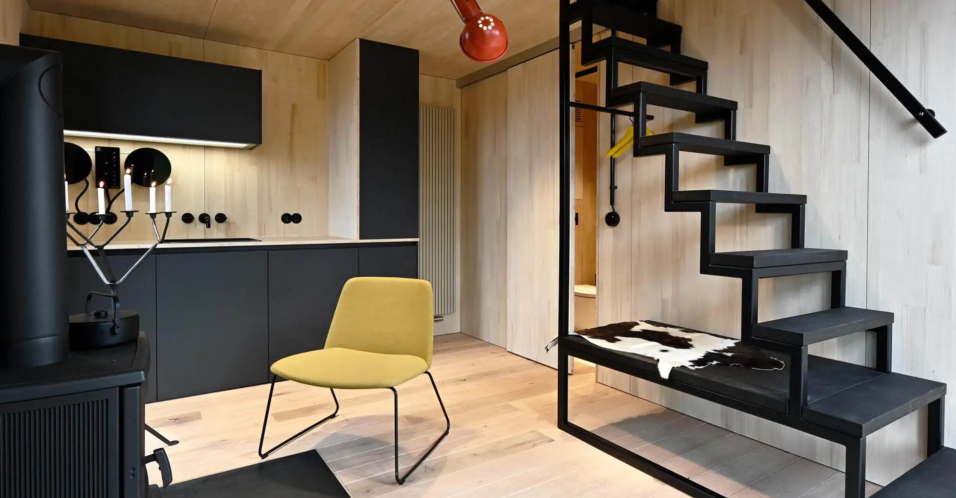Kompaktes Wohninterieur mit schwarzer Stahlwangentreppe, gelbem Sessel und Holzoberflächen.