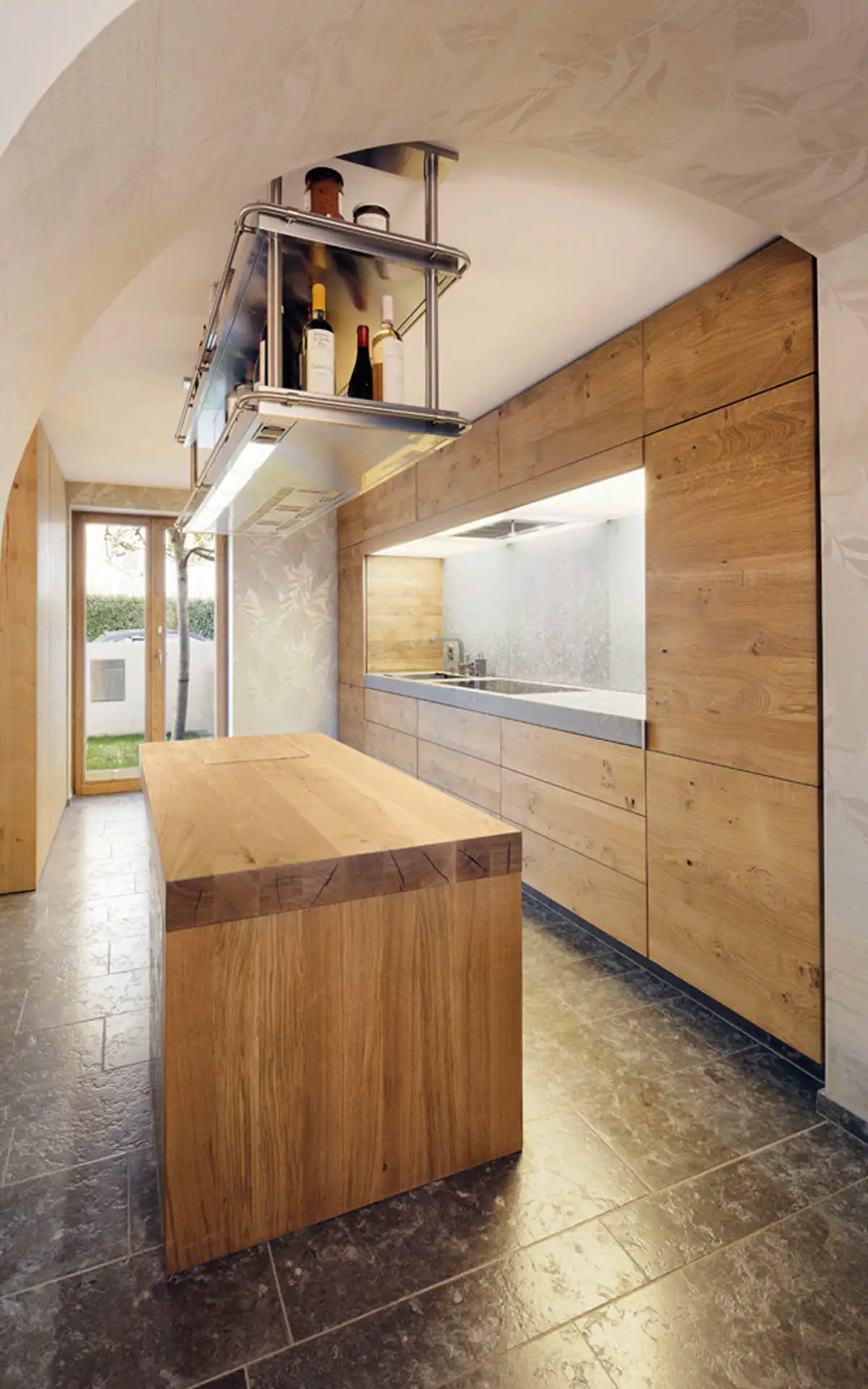 Schmale Küche unter Gewölbe: durchgehende Eichenfront, freistehende Arbeitsinsel und deckenmontiertes Edelstahlregal.