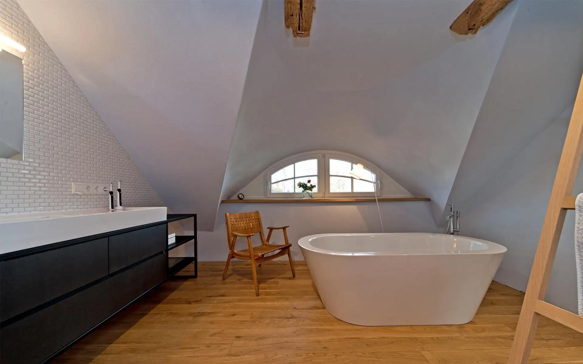 Bad unterm Satteldach: freistehende Wanne vor Bogenfenster, Holzfußboden, schwarzer Waschtisch mit weißer Auflage.