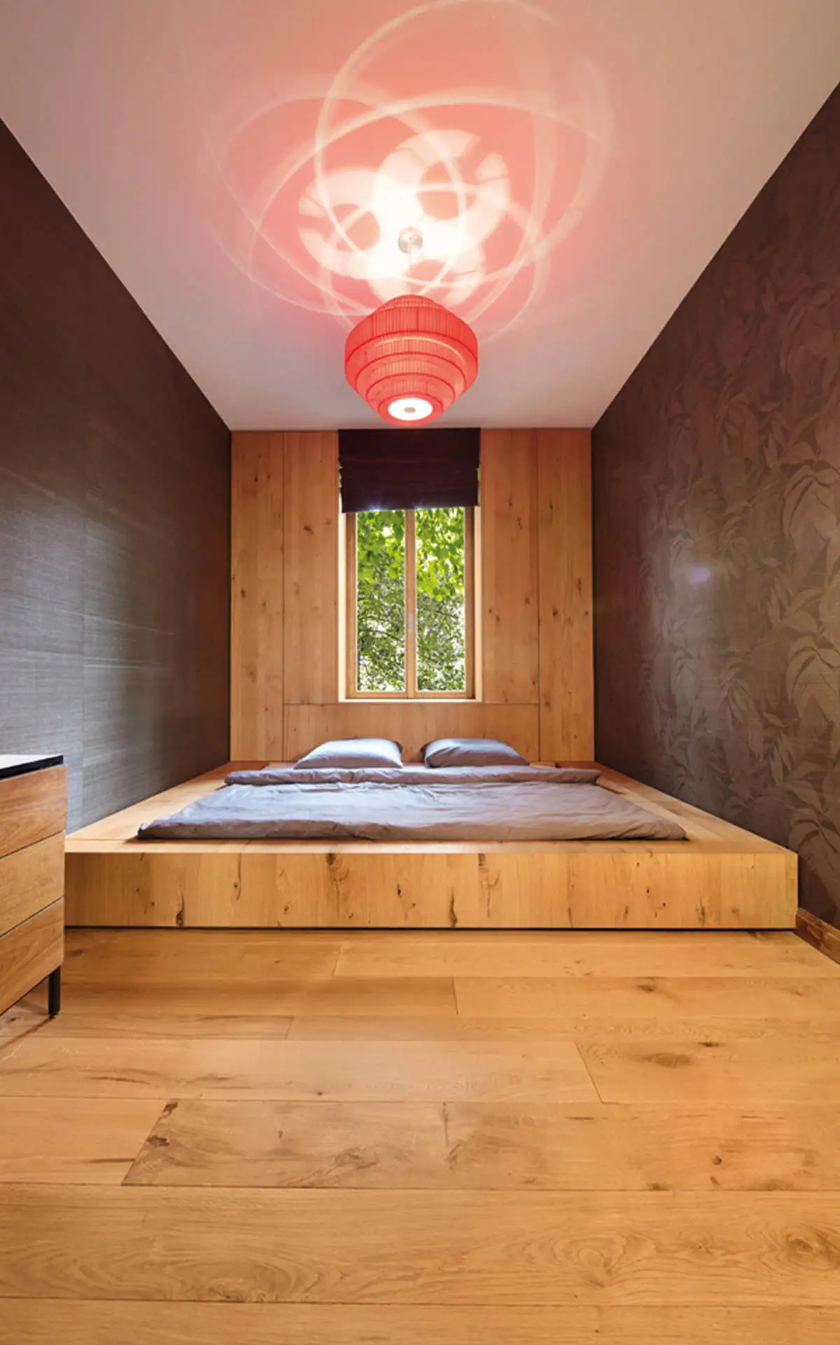 Schlafzimmer mit bodennahem Podestbett aus Eiche; dunkle Wandbespannung und rote Deckenleuchte mit Lichtreflexen.