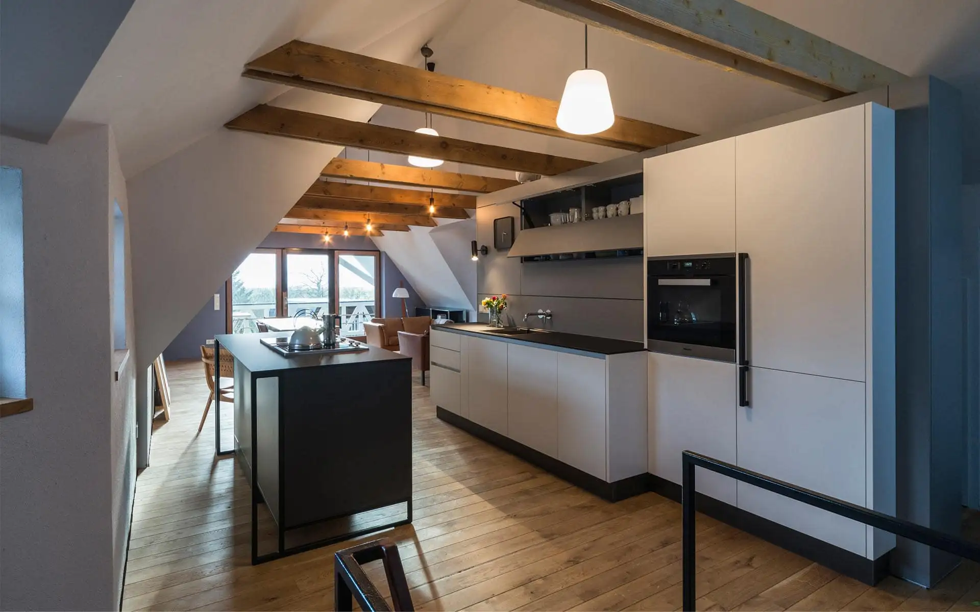 Offene Küche im Dachgeschoss mit Balkendecke, dunkler Insel und Blick zur Terrasse im Hintergrund.