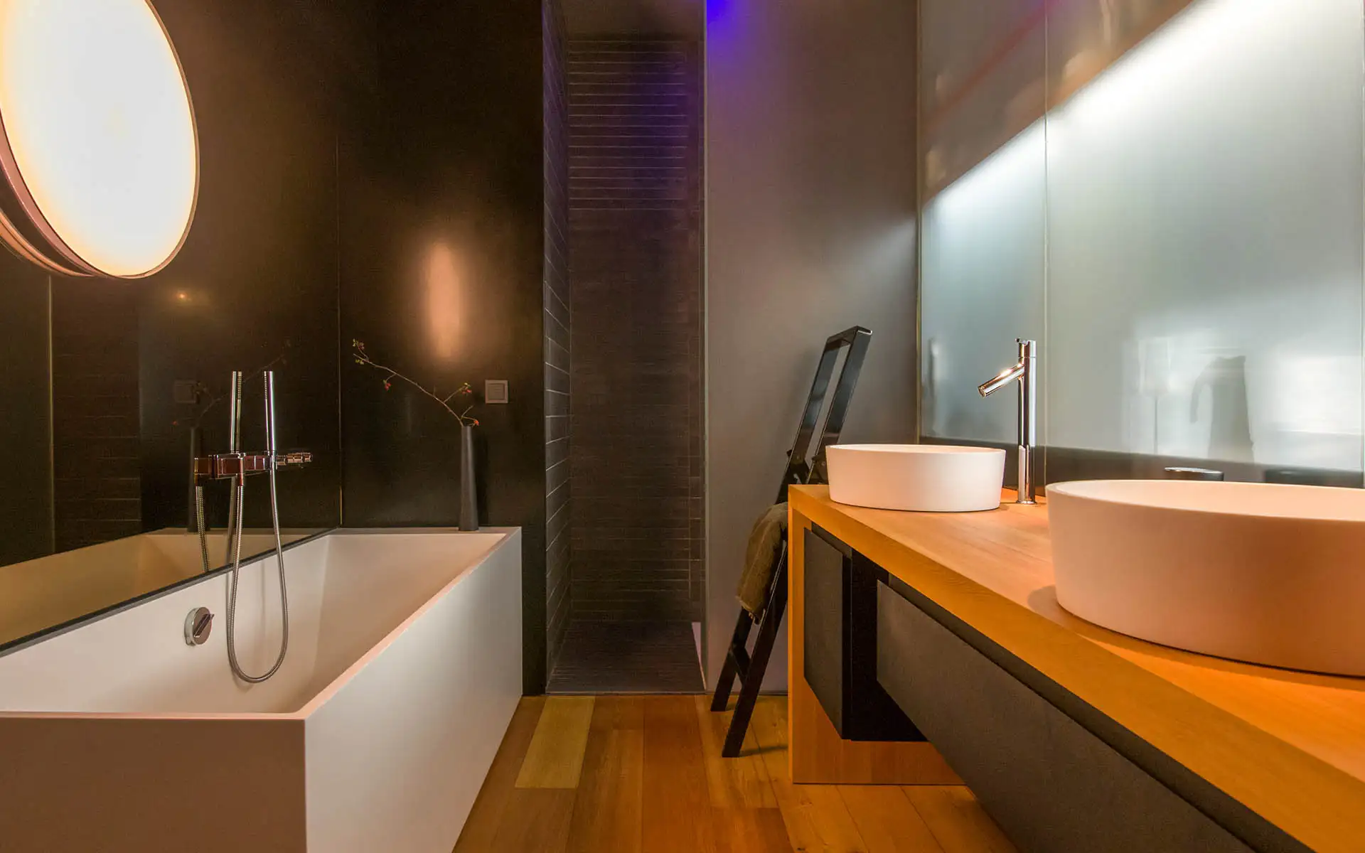 Bad mit weißer Wanne, Doppelwaschplatz auf Holzplatte und dunklen Wandflächen; runde Leuchte links.