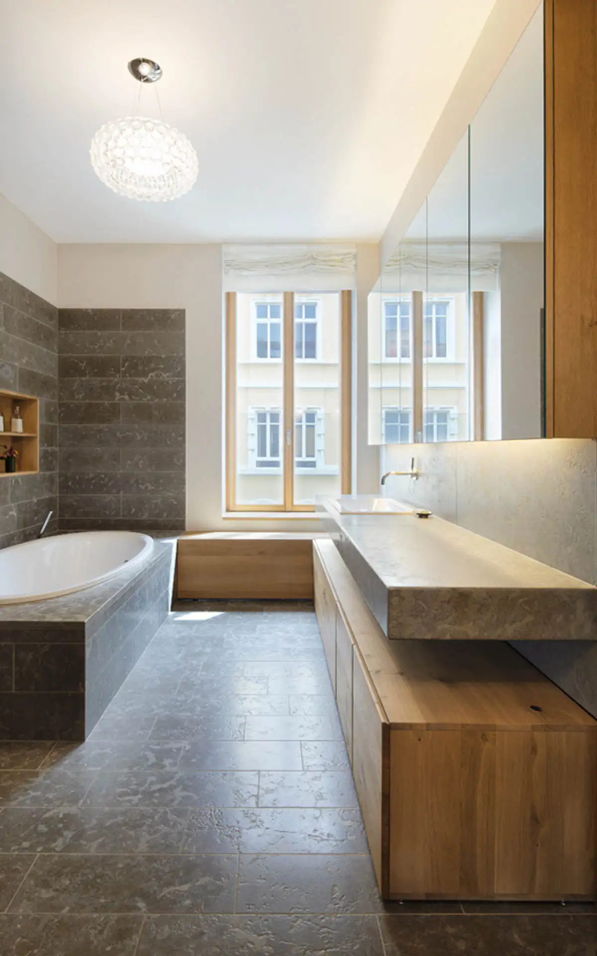 Bad mit halbrunder Wanne, grauen Steinfliesen, wandlangem Waschtisch mit Steinauflage und Spiegelschränken vor Fensterfront.